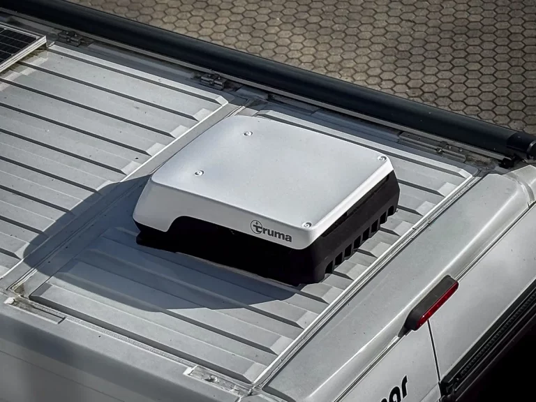 Close-up of a silver Truma appliance mounted on a metal truck bed, with vent slots visible and a black rail nearby.