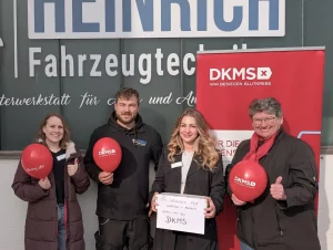Group of four people smiling and posing with red DKMS balloons in front of a vehicle workshop sign reading 'Fahrzeugtechnik'.